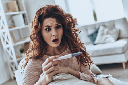 No way! Terrified young women looking at pregnancy test and keeping mouth open while sitting on the sofa at home        の写真素材