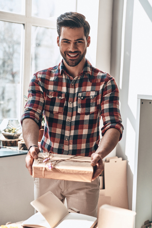 With love for you. Handsome young man giving a gift box and looking at camera with smile while standing in the creative working spaceの写真素材