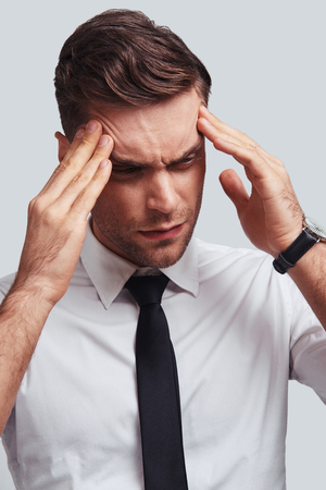 Terrible headache. Frustrated young man in full suit touching his head with hands while standing against grey backgroundの写真素材