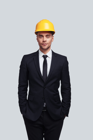 Safety comes first. Good looking young man in hardhat keeping hands in pockets and looking at camera while standing against grey backgroundの写真素材
