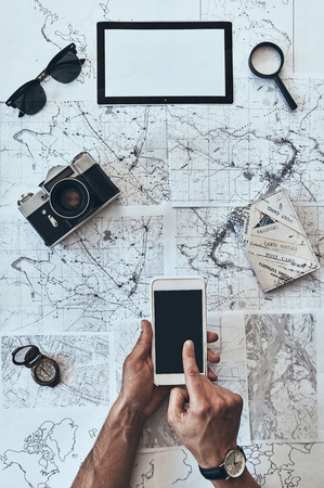 New directions. Close up top view of man using smart phone with sunglasses, photo camera, compass, magnifying glass and passport lying on map aroundの写真素材