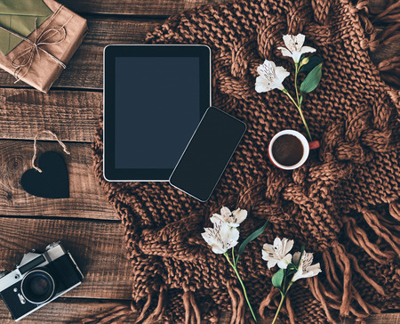 Modern technologies. High angle shot of knitted scarf, flowers, cup of coffee, gift box, photo camera, smart phone and digital table on wooden deskの写真素材