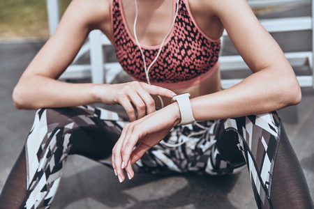 What time is it? Close up of young woman in sports clothing checking the time while sitting outdoorsの写真素材