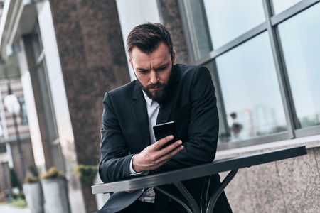 Taking everything seriously. Good looking young man in full suit using his smart phone while sitting in the cafe outdoorsの写真素材