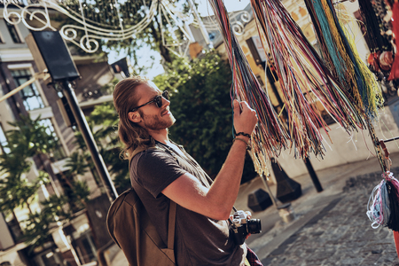 What to choose? Handsome young man in casual clothing choosing necklace and smiling while standing near the market stall outdoorsの写真素材