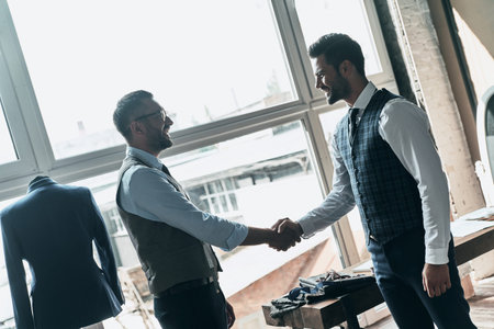 elcome! Two young fashionable men shaking hands and smiling while standing in workshopの写真素材