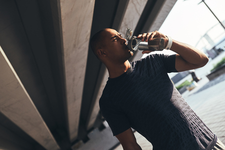 Hydrating. Young African man in sports clothing drinking water while standing outsideの写真素材