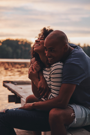 Too good to be true. Happy young couple embracing and smiling while sitting on the pier near the lakeの写真素材