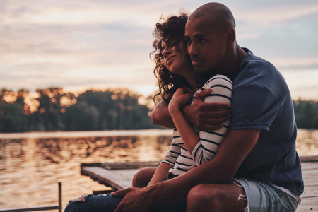 Romantic couple. Happy young couple embracing and smiling while sitting on the pier near the lakeの写真素材