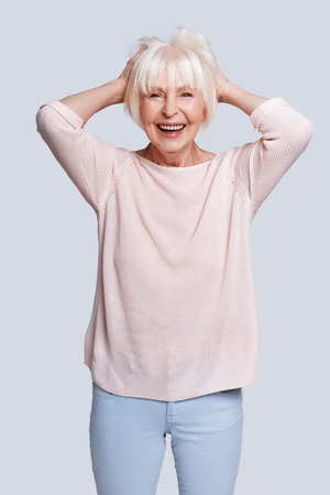No cares. Beautiful senior woman keeping hands in hair and smiling while standing against grey backgroundの写真素材