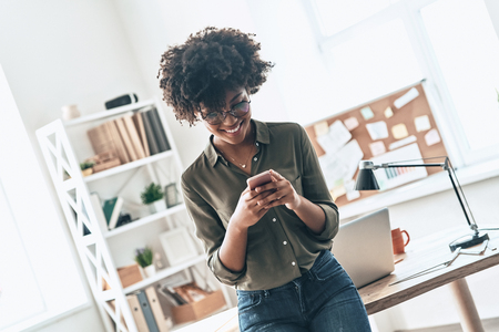 Chatting with client. Attractive young African woman using her smart phone and smiling while standing in the officeの写真素材