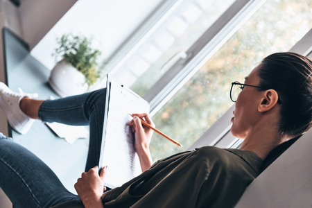 Thinking about every detail. Top view of concentrated young woman writing something down while sitting on the window sill in the officeの写真素材
