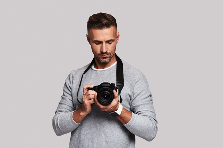 Confident photographer. Good looking young man holding digital camera while standing against grey backgroundの写真素材