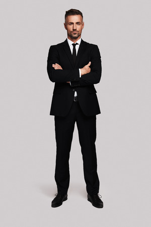 Young and successful. Full length of handsome young man in full suit keeping arms crossed and looking at camera while standing against grey backgroundの写真素材
