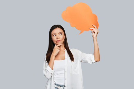 What if? Beautiful young Asian woman keeping hand on chin while standing against grey backgroundの写真素材