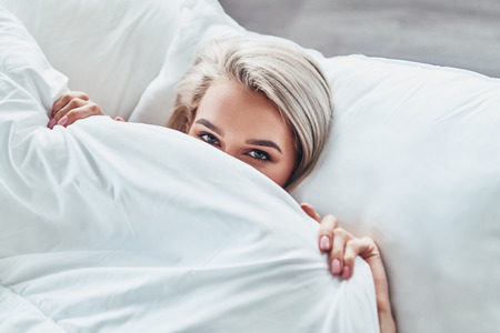 Too lazy to wake up. Top view of attractive young woman covering half of her face with blanket and looking at camera while lying in bed at homeの写真素材