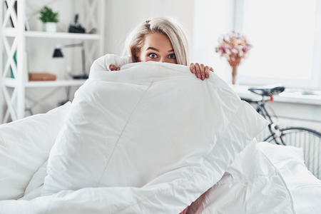 Monday morning. Attractive young woman covering half of her face with blanket and looking at camera while sitting in bed at homeの写真素材