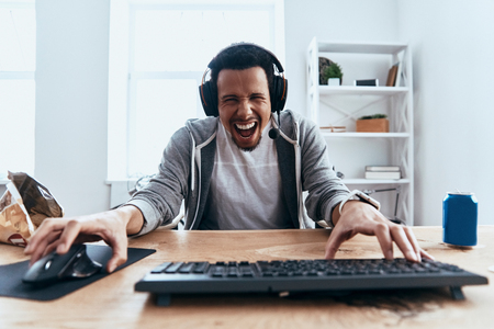 Carefree and happy. Handsome young man in casual clothing looking at camera and smiling while playing computer games at homeの写真素材