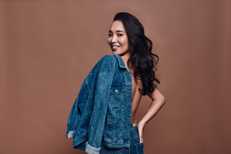 Pure feminine beauty. Attractive young woman carrying on shoulders denim jacket and smiling while standing against grey backgroundの写真素材