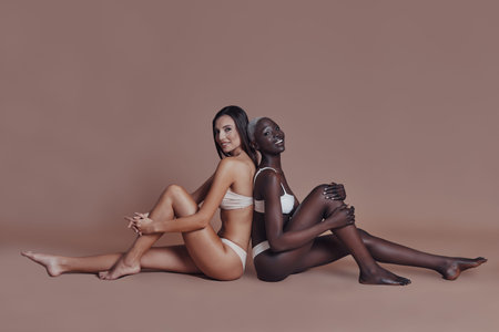 Feeling free to express sensuality. Two attractive mixed race women looking at camera while sitting back to back against brown backgroundの写真素材