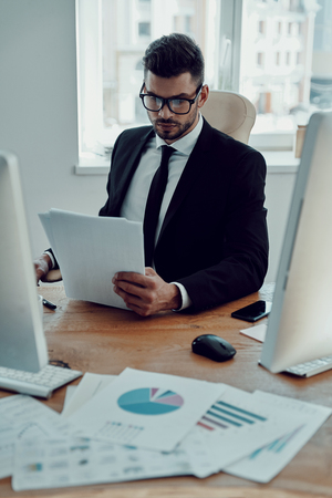 Checking every detail. Serious young man in formalwear reading contract while sitting in the officeの写真素材