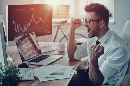 Unbelievable growth. Happy young businessman in formalwear shouting and gesturing while using computer in the officeの写真素材