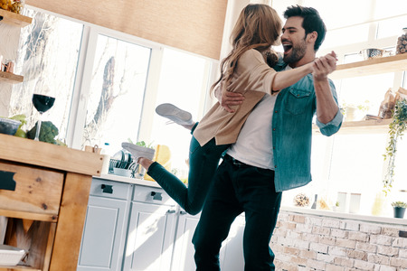 So great to be together! Full length of beautiful young couple in casual clothing dancing and smiling while standing in the kitchen at homeの写真素材