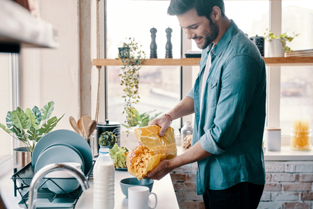 Easy breakfast. Handsome young man in casual wear preparing corn flakes and smiling while standing in the kitchen at homeの写真素材