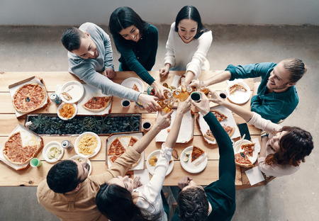 Cheers! Top view of young people in casual wear toasting each other and smiling while having a dinner party indoorsの写真素材