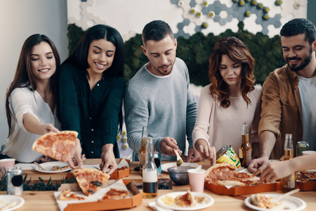 Choosing the best slice. Group of young people in casual wear eating pizza and smiling while having a dinner party indoorsの写真素材