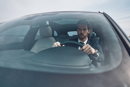 Busy day. Handsome young man in full suit looking straight while driving a carの写真素材