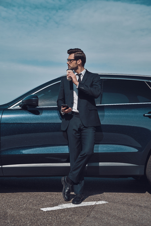 Time for a little break. Full length of handsome young businessman using smart phone and drinking coffee while standing near his car outdoorsの写真素材