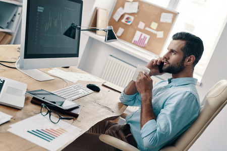 Talking about business. Top view of young modern businessman talking on the phone and analyzing data using computer while sitting in the officeの写真素材