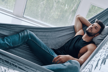 Simply resting. Top view of handsome young man in casual clothing relaxing while lying in the hammock indoorsの写真素材