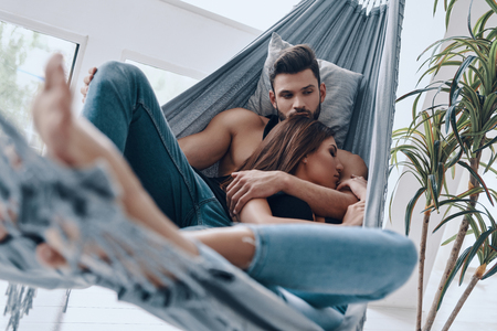 Carefree day together. Beautiful young couple embracing while resting in the hammock indoorsの写真素材