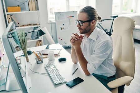 Concidering next step. Top view of thoughtful young man in shirt working using computer while sitting in the officeの写真素材