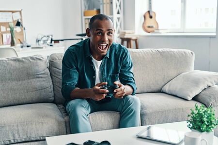 So much fun! Handsome young African man playing video game and smiling while sitting indoorsの写真素材