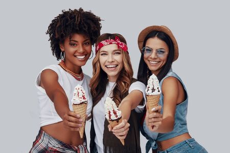 Three attractive young women eating ice cream and smiling while standing against grey backgroundの写真素材