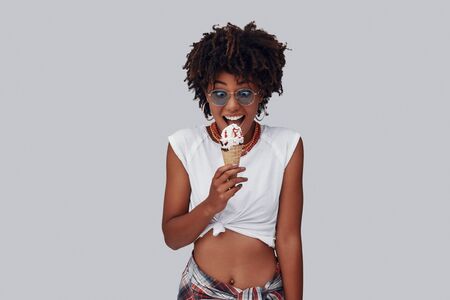 Attractive young African woman eating ice cream and smiling while standing against grey backgroundの写真素材