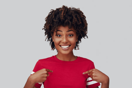 Attractive young African woman looking at camera with smile and pointing at herself while standing...
