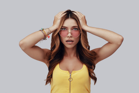 Surprised young woman looking at camera and keeping head in hands while standing against grey...