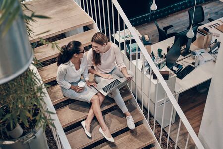 Top view of two young modern women in smart casual wear communicating and using laptop while sitting on the stairsの写真素材