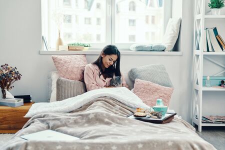 Beautiful young woman in pajamas smiling and embracing domestic cat while resting in bed at home                   の写真素材