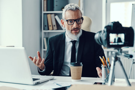 Confident mature man in elegant suit telling something and gesturing while making social media video