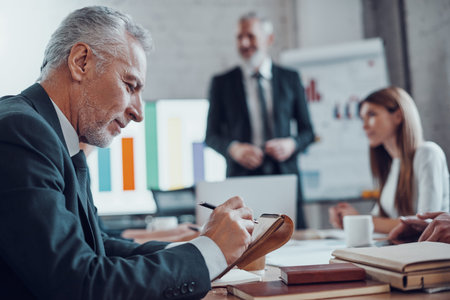 Mature businessman writing something down and smiling while working together with colleagues in the...