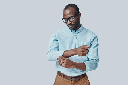 Fashionable young African man adjusting sleeve while standing against grey background