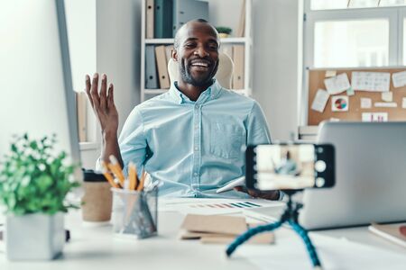 Handsome young African man in shirt telling something and smiling while making social media videoの写真素材