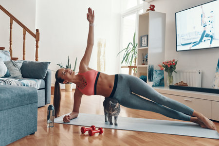 Happy young woman in sports clothing standing in plank position with cat walking around while...
