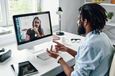 Busy young African man in shirt talking with his coworker using computer while sitting indoorsの写真素材