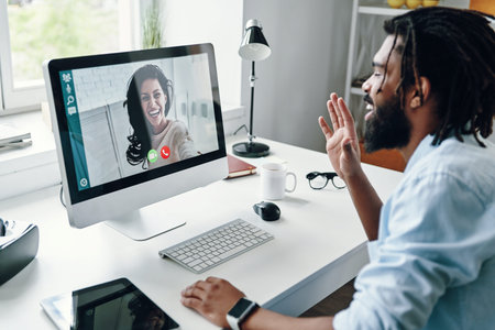 Confident young African man in shirt talking with smiling woman using computer while sitting indoors